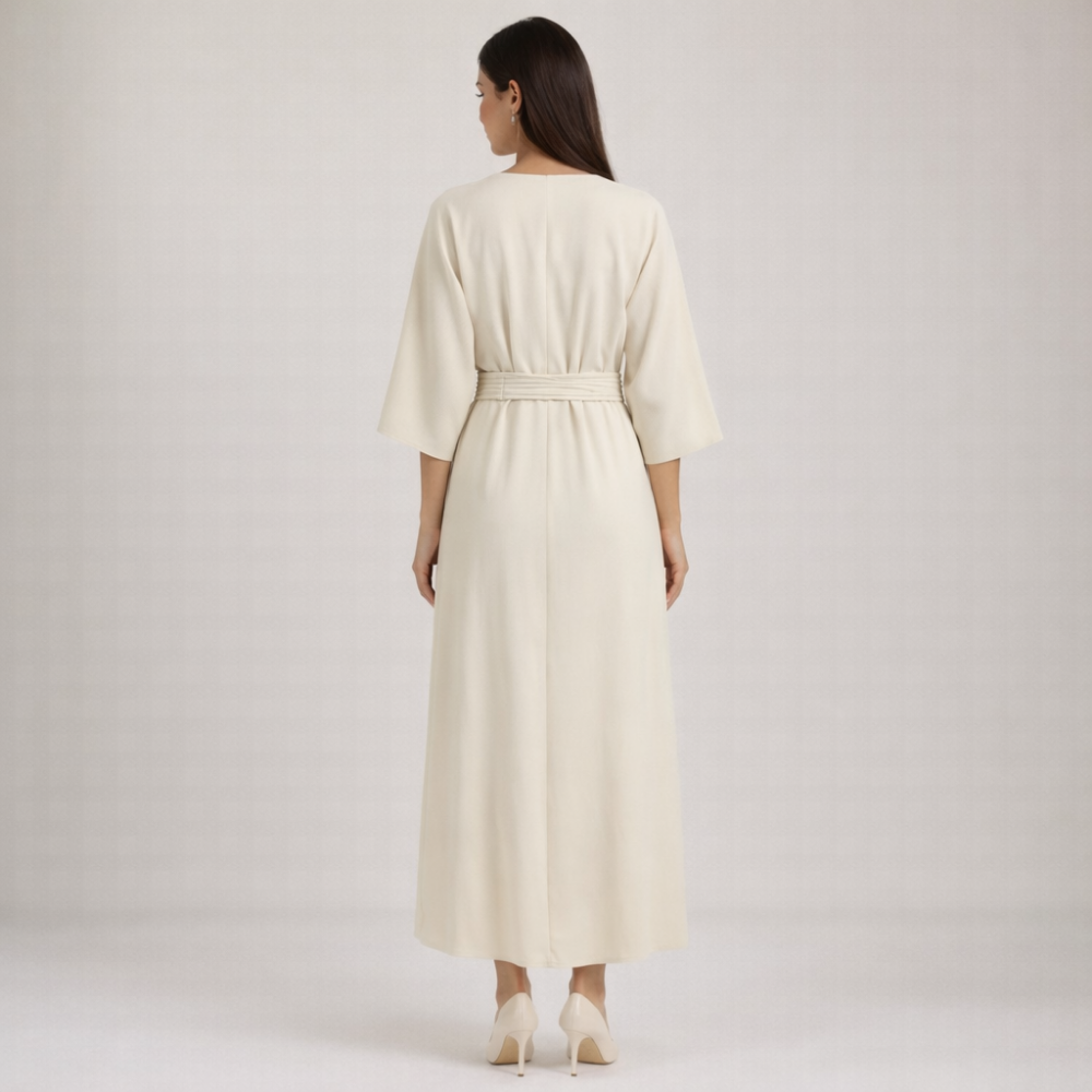 A woman in the Evergreen Outfits Maxi Dress Women Belted Waist With Bell Shaped Sleeves stands with her long brown hair over the cream dress, featuring wide sleeves and a belted waist, paired with matching heels in a minimal, light-toned space.